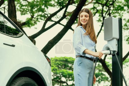 Photo for Young woman recharging battery for electric car during road trip travel EV car in natural forest or national park. Eco friendly travel during vacation and holiday. Exalt - Royalty Free Image
