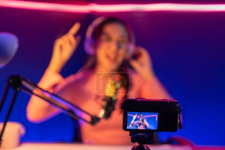 Photo for Selective focus camera screen in host channel of beautiful Asian listening the music on social media with listener, wearing pastel headphones on blurred background using mic at neon studio. Stratagem. - Royalty Free Image