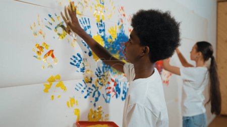 Foto de Vista posterior del niño africano feliz pintado la pared manchada con la mano colorida mientras que usa la camisa blanca casual en la lección de arte.Estudiante del mercado utiliza la impresión de la mano para hacer ilustraciones creativas. Educación. Edificación. - Imagen libre de derechos