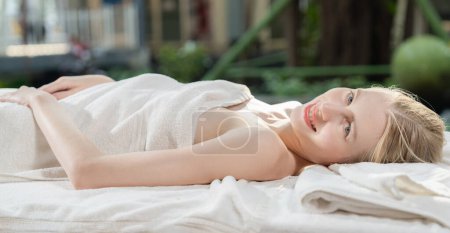 Photo for Beautiful young woman relaxes on a spa bed surrounded by nature. ready for a body massage. Attractive female in white towel lying peacefully during waiting for body massage. Close up. Tranquility - Royalty Free Image