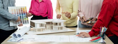 Zugeschnittenes Bild eines professionellen Architekten-Teams Brainstorming über die Farbauswahl des Hauses, während der Architekt auf einem Blaupause Dokument am Tisch mit Blaupause und Hausmodell platziert schreibt. Bunt gemischt.