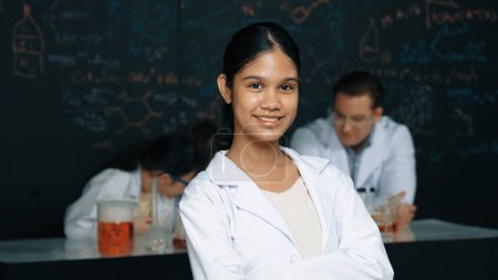 Photo for Girl looking at camera with arm folded while people doing experiment at laboratory. Cute student standing blackboard with chemical theory with blurring background at STEM science class. Edification. - Royalty Free Image