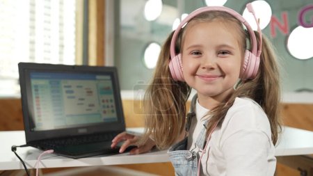 Smiling smart caucasian girl show system programing or coding program in STEM technology education. Child wearing headphone while working by using laptop writing code and look at camera. Erudition.
