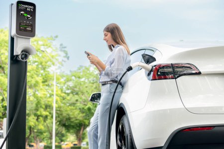 Photo for Young woman recharge EV electric vehicle battery from EV charging station and using smartphone online banking to pay for electricity in city park. Eco friendly vehicle travel with EV car. Exalt - Royalty Free Image