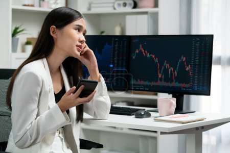 Stressful young beautiful Asian businesswoman focusing on smartphone, analyzing possible risk to profit or loss in dynamic exchange stock market graph on pc screen at modern office. Stratagem.