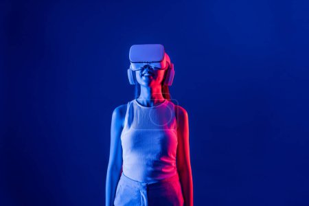 Foto de Soporte femenino inteligente con auriculares VR rodeados de luz de neón cyberpunk que conectan tecnología de comunidad ciberespacial metaversa y futurista. Mujer elegante mirada generada área virtual. Alucinaciones. - Imagen libre de derechos