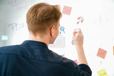 Photo for Professional caucasian businessman writing and sharing marketing idea by using mind map and sticky notes on glass board at meeting room. Creative business and planing concept. Back view. Immaculate. - Royalty Free Image