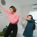 Happy attractive mother dancing to music together with asian daughter at bedroom. Caucasian mom dance or exercise while cute child move to music while jump to bed. Family recreation concept. Pedagogy.