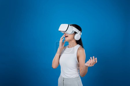 Foto de Mujer inteligente de pie con fondo azul con auriculares VR que conectan tecnología de comunidad ciberespacial metaversa y futurista. Mujer elegante emocionado de ver generado paisaje virtual. Alucinaciones. - Imagen libre de derechos
