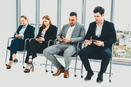 Foto de Empresarias y hombres de negocios que utilizan el teléfono móvil mientras esperan en sillas en la oficina para la entrevista de trabajo. Negocios corporativos y concepto de recursos humanos. BARROS - Imagen libre de derechos