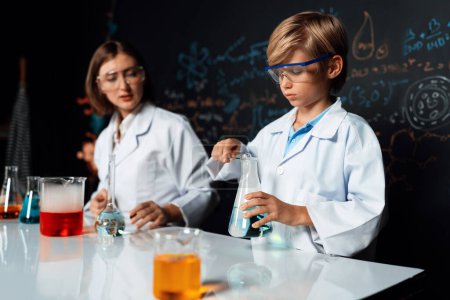Photo for Teacher support schoolboy in laboratory they wear lab coat and glasses stand and experiment about science of chemistry in STEM class. Student tong solid down to yellow liquid in beaker. Erudition. - Royalty Free Image
