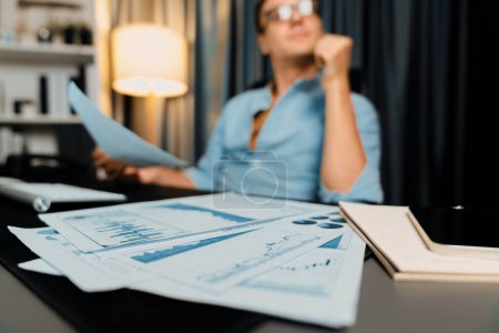 Photo for Blurred of photo with smart businessman looking on business paragraph dynamic data marketing analysis planning sheet surrounded paper sheet and pc monitor report at home office at night. Pecuniary. - Royalty Free Image