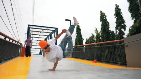 Photo for Panorama shot of professional hipster perform b-boy performance and energetic footstep. Skilled happy man practice break dance while friend cheering him at bridge. Outdoor sport 2024. Sprightly. - Royalty Free Image