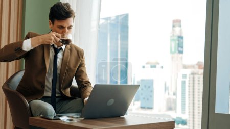 Photo for Businessman sitting on furniture working on laptop at ornamented corporate waiting area with cityscape background on the window. Business profession and strategic marketing plan for business success. - Royalty Free Image