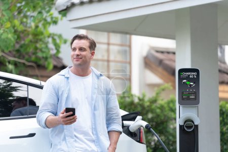 Photo for Modern eco-friendly man recharging electric vehicle from home EV charging station. Innovative EV technology utilization for tracking energy usage to optimize battery charging at home. Synchronos - Royalty Free Image