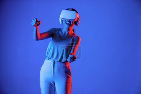 Photo for Smart female stand surrounded by neon light wearing VR headset connecting metaverse, future cyberspace community technology. Elegant woman enjoy playing car racing games in meta world. Hallucination. - Royalty Free Image