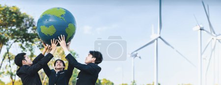 Foto de Concepto del Día de la Tierra con gran globo terráqueo sostenido por el equipo de empresarios asiáticos promoviendo la conciencia ambiental utilizando energía limpia sostenible y renovable con turbina eólica para un futuro más verde. Gyre. - Imagen libre de derechos