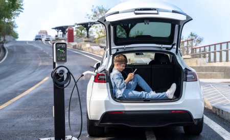 Photo for Little boy sitting on car trunk, using smartphone while recharging eco-friendly car from EV charging station. EV car road trip travel as alternative vehicle using sustainable energy concept. Perpetual - Royalty Free Image