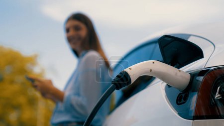 Photo for Woman recharging battery for electric car during road trip travel EV car in autumanl season natural forest or national park. Eco friendly travel during vacation and holiday. Exalt - Royalty Free Image