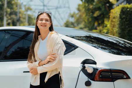 Téléchargez les photos : Jeune femme recharger son véhicule électrique EV au parking vert du parc de la ville. Style de vie urbain durable pour voiture écologique EV avec station de charge de la batterie. Expedient - en image libre de droit