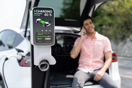 Photo for EV car recharging battery from outdoor EV charger display battery status on blur background of man using smartphone with natural scenic as concept of eco-friendly travel with clean energy. Perpetual - Royalty Free Image
