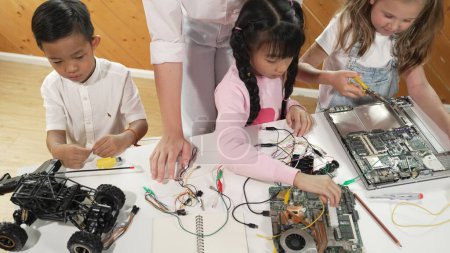 Draufsicht auf einen kaukasischen Lehrer, der diverse Schüler über elektronische Tafeln unterrichtet. Multikulturelle Kinder lernen durch den Einsatz von Chips und Drähten über digitales elektrisches Werkzeug und die Befestigung von Motherboards. Erution.