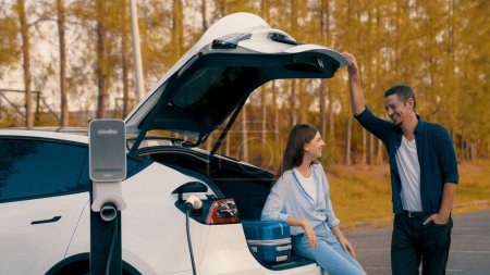 Das Paar tankt sein Elektroauto mit einer Ladestation an der Raststätte während der herbstlichen Fahrt in den Nationalpark. Erholsame Ferien mit umweltfreundlichem Auto. Erhöhen