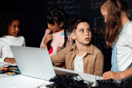 Photo for In classroom student in STEM class learning about coding robotics car. Brown shirt Schoolboy and white bib schoolgirl watching laptop and discussing. Other schoolgirls playing around funny. Erudition. - Royalty Free Image