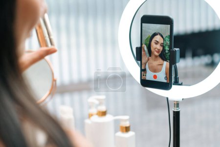Rear view young woman making beauty and cosmetic tutorial video content for social media. Beauty blogger showing how to apply beauty care to audience or follower on camera screen. Blithe