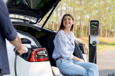 Photo for Lovely young couple recharging battery for electric car while relaxing and sitting on the trunk during road trip travel EV car in autumnal forest. Eco friendly travel on vacation during autumn. Exalt - Royalty Free Image