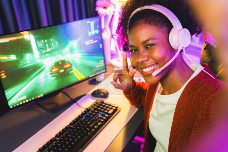 Photo for Host channel African of gaming streamer, African girl playing car racing game with joystick, wearing pastel headsets with mic, looking at the camera against background in neon color room. Tastemaker. - Royalty Free Image