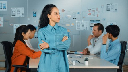 Hispanic professional businesswoman standing while looking at camera in creative business meeting. Beautiful confident leader posing while diverse business people discussion about plan. Manipulator.