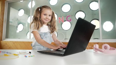 Foto de Pequeña niña linda usando el ordenador portátil para trabajar o jugar juegos. Estudiante trabajando en la computadora mientras mira y sonríe a la cámara. Los niños caucásicos asisten a clases en línea usando auriculares. Erudición. - Imagen libre de derechos