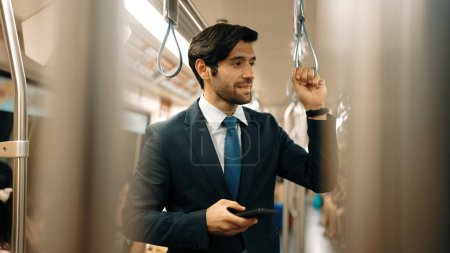 Téléchargez les photos : Homme d'affaires compétent debout dans le train ou le métro tout en tenant le téléphone. Gestionnaire de projet professionnel regardant autour tout en profiter en train tout en jouant téléphone mobile à la gare. Exultant. - en image libre de droit