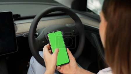 Photo for Holiday vacation road trip with environmental-friendly car concept. Eco-conscious woman on driver seat holding blank copyspace green screen smartphone for EV battery status. Exalt - Royalty Free Image
