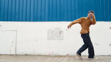 Foto de El hombre hispano estira los brazos y baila en la calle bailando frente a la pared. Motion shot de bailarina con estilo o coreógrafa en traje casual que practica el baile en estilo hip hop. Deporte al aire libre 2024. Endeavor. - Imagen libre de derechos