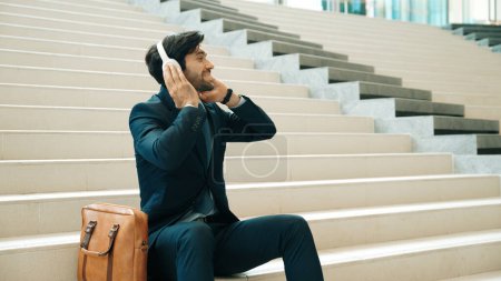 Téléchargez les photos : Homme d'affaires professionnel cueillette et écoute de la musique en utilisant un casque tout en étant assis à l'escalier. Chef de projet danse tout en écoutant la chanson tout en portant un casque, costume. Fond blanc. Exultant. - en image libre de droit