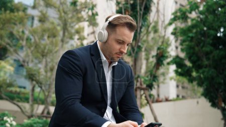 Business people wear his headphone to listen relaxing music while using phone open song or chatting with marketing team in green city. Happy project manager move to song while hold telephone. Urbane.