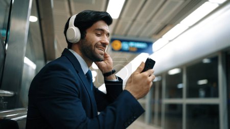 Téléchargez les photos : Homme d'affaires intelligent écoutant de la musique par casque tout en attendant train à la gare avec fond flou. Gestionnaire de projet compétent profiter écouter un son relaxant tout en tenant le téléphone mobile. Exultant. - en image libre de droit