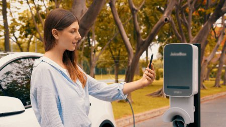 Femme utilisant smartphone application bancaire en ligne pour payer la charge de la batterie de voiture électrique à partir de la station de charge EV pendant les vacances voyage sur la route dans le parc national ou la forêt d'été. Exalt.