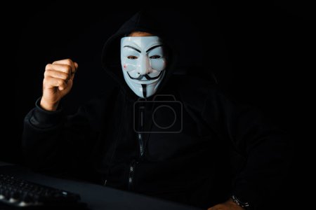 Photo for White smiling criminal anonymous mask looking at camera with raising fist up, installing virus encryption through main database server system in succeed at privacy on computer ransom company. Surmise. - Royalty Free Image