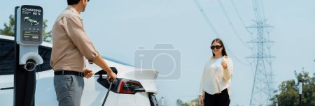 Photo for Young couple recharge EV car battery at charging station connected to power grid tower electrical industrial facility as electrical industry for eco friendly vehicle utilization. Panorama Expedient - Royalty Free Image
