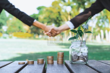 Sustainable money growth investment, glass jar filled with money saving and coin stack with businesspeople handshake in background as eco-friendly financial investment nurtured with nature. Gyre