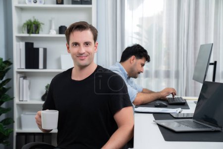 Téléchargez les photos : Profil de souriant pigiste ou homme d'affaires regardant caméra avec tasse de café tout collègue travaillant sur ordinateur portable avec de la paperasserie au bureau moderne. Thème du projet créatif sur le lieu de travail. Vendu. - en image libre de droit