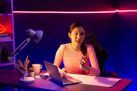 Photo for Smiling creative young beautiful Asian influencer talking in positive podcast live on social media speech at night, using laptop for presentation online at pink neon light modern studio. Stratagem. - Royalty Free Image