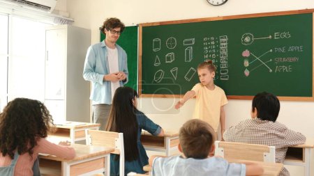 Kluger Junge zeigt auf einen Freund, während attraktive Kinder im Unterricht sitzen. Grundschüler reden und erklären dem kaukasischen Lehrer, während sie vor der Klasse stehen und zuhören. Pädagogik.