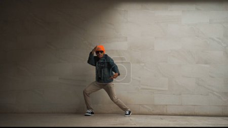 Foto de Motion shot of hipster realizar break dance en el fondo gris. Bailarina callejera practica el movimiento hip hop mientras usa un elegante paño de hip hop y se mueve a la música funky. Deporte al aire libre 2024. Rígidamente.. - Imagen libre de derechos