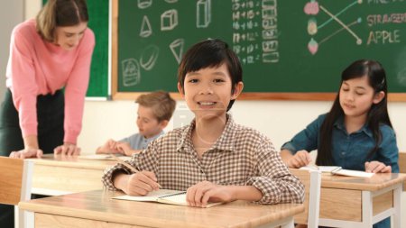 Photo for Asian smart boy smiling to camera while student writing answer in answer sheet. Multicultural student doing classwork or test while caucasian teacher checking student homework at classroom. Pedagogy. - Royalty Free Image