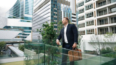 Caucasian business man hold mobile phone and looking at skyscraper view. Skilled male leader standing rooftop in modern building. Caucasian executive manager look at phone while rest his eyes. Urbane.
