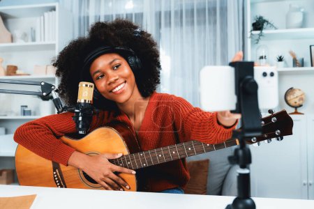 Téléchargez les photos : Canal hôte de belle femme africaine influenceur réglage smartphone, chanter avec jouer de la guitare en diffusion. créneau horaire du blogueur de musique sur les médias sociaux en direct en ligne. Concept de créateur audio. Tastemaker. - en image libre de droit